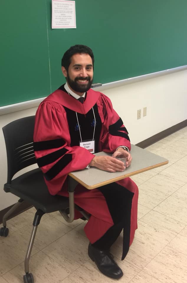 Male student wearing black and red graduation gown sitting at desk with glass of water