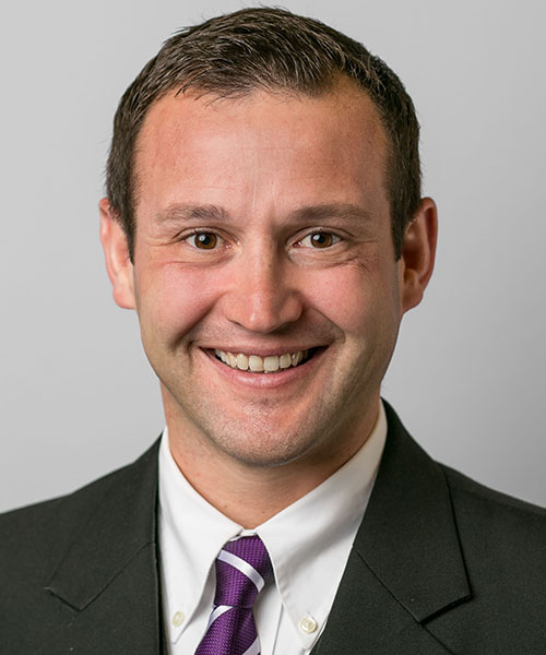 Recent alumnus Anthony DeMattee poses against a gray background, dressed in a dark suit and tie.