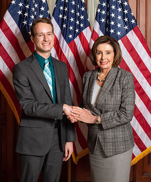 A Political Science undergraduate student poses with House Speaker Nancy Pelosi in front of three American flags.