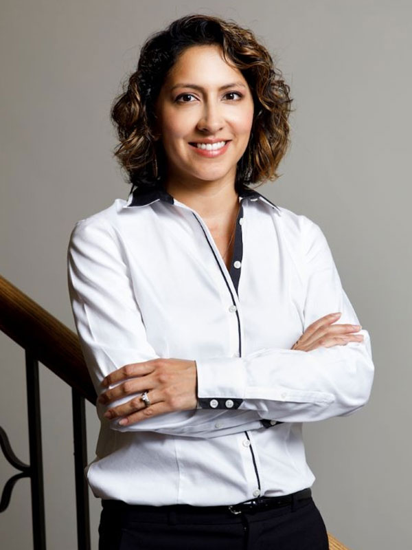 A headshot of Vanessa Cruz-Nichols, who wears a white buttoned shirt and poses on a staircase.