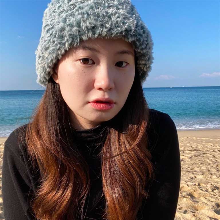 A headshot of Joohyun Kim, who wears a black shirt and poses on a beach with the ocean in the background.