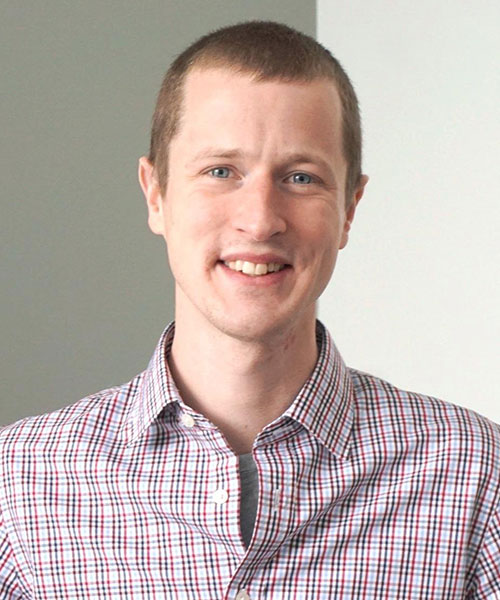 Faculty member Steven Webster poses against a white and gray background, dressed in a checkered button-up shirt.